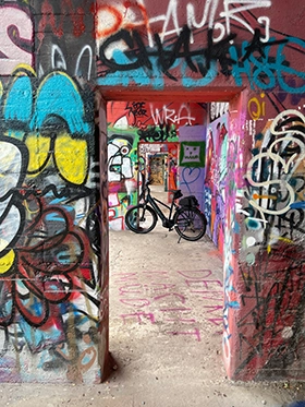 nnenraum einer betonierten, vollständig mit bunten Graffiti bedeckten Ruine; mehrere hintereinander liegende Türöffnungen bilden einen tiefen Blick, in der Mitte steht ein schwarzes Fahrrad.