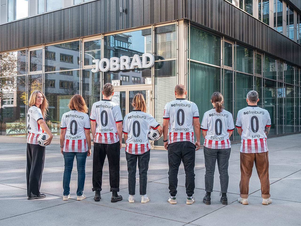 Mitarbeitende von JobRad stehen aufgereiht mit SC Trikots auf dem Campus, sie stehen mit dem Rücken zu uns und bilden 2000000 mit ihrer Trikotrückseite. Mitarbeitende von JobRad stehen aufgereiht mit SC Trikots auf dem Campus, sie stehen mit dem Rücken zu uns und bilden 2000000 mit ihrer Trikotrückseite.